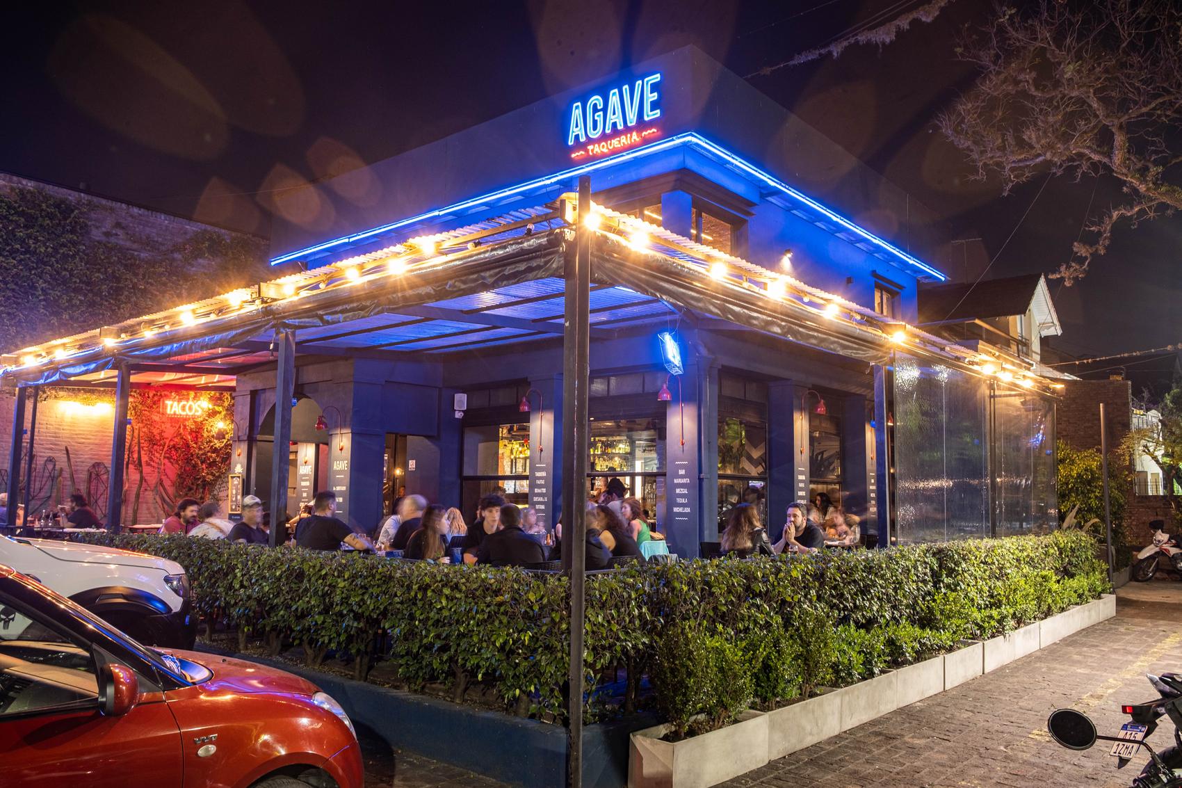 Frente de AGAVE de noche, con el cartel de neón azul y la terraza llena de gente